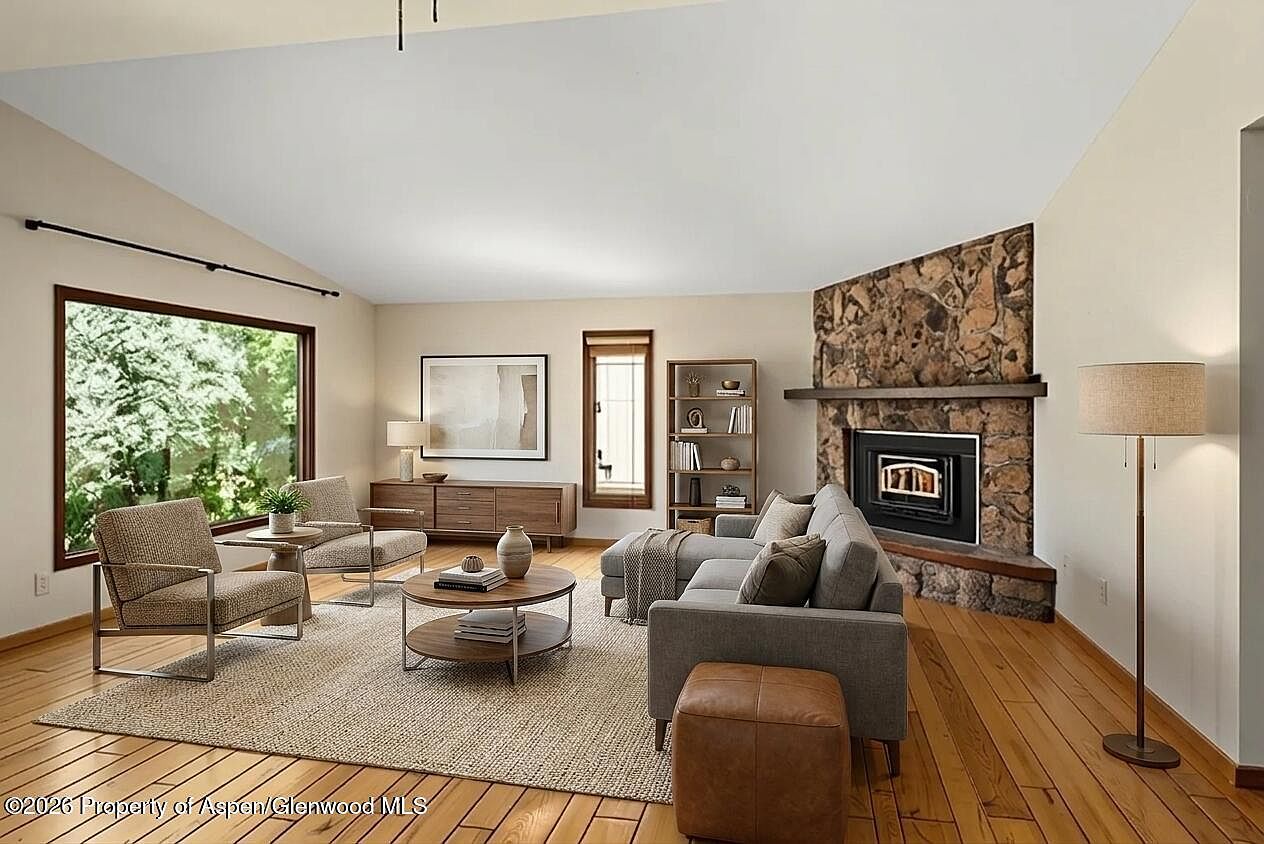Vaulted great room with floor-to-ceiling river-stone fireplace, wood floors, sectional seating, and large picture window to the side garden