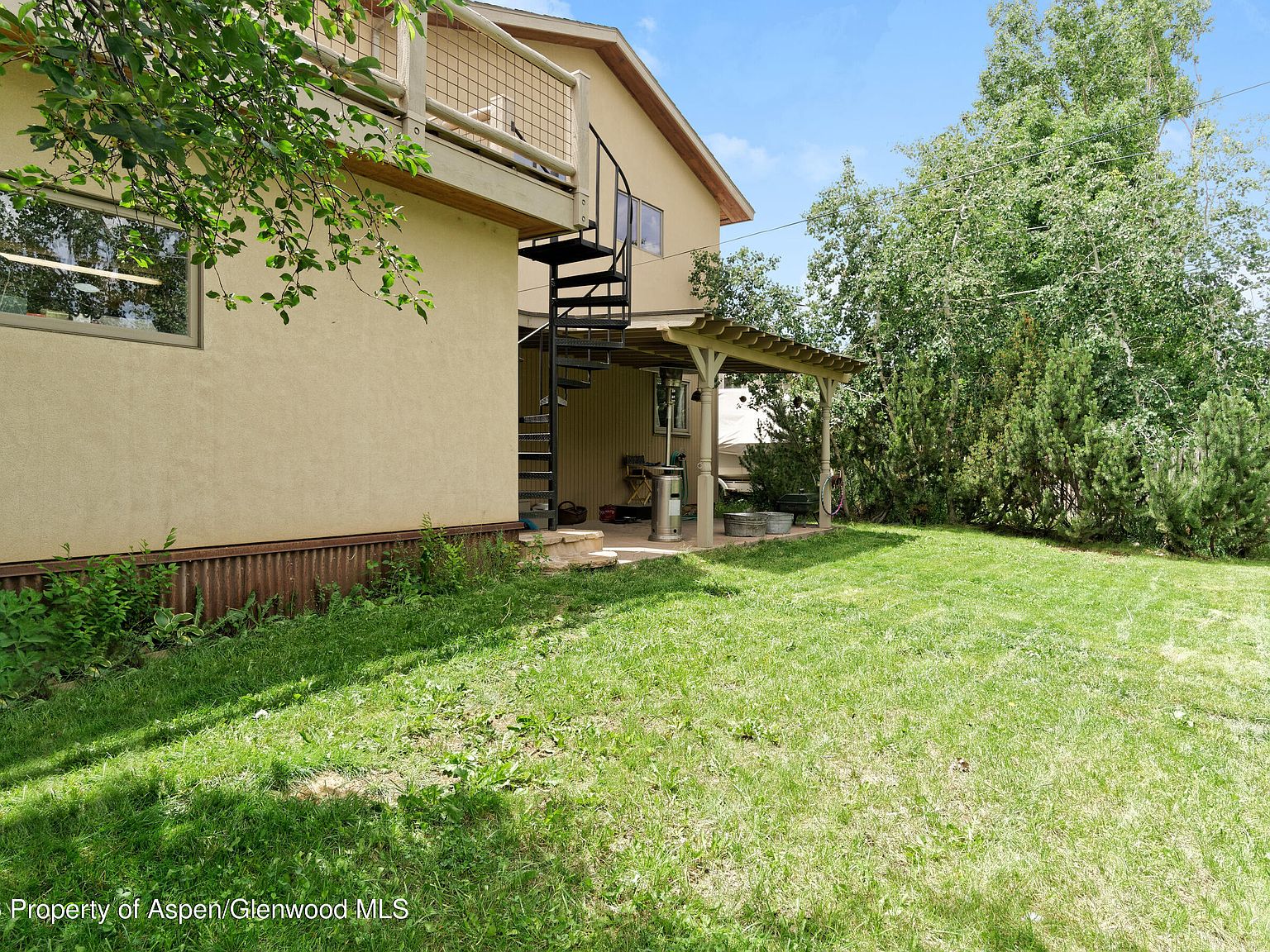 Back exterior of the home with two-story stucco facade, second-floor deck off the primary suite, spiral staircase to ground, and a generous fenced lawn bordered by mature cottonwoods